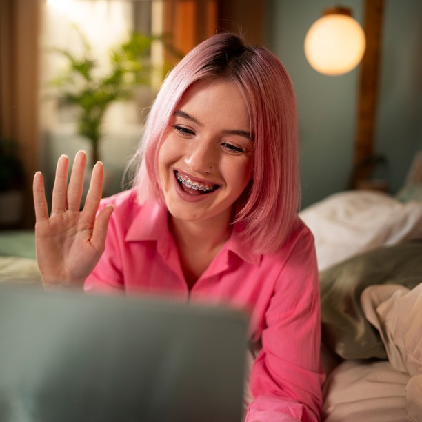A girl with pink hair and braces waves while on a video call.