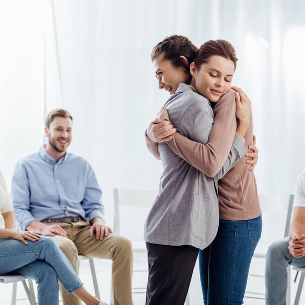 Two women share a supportive hug during a group therapy session.